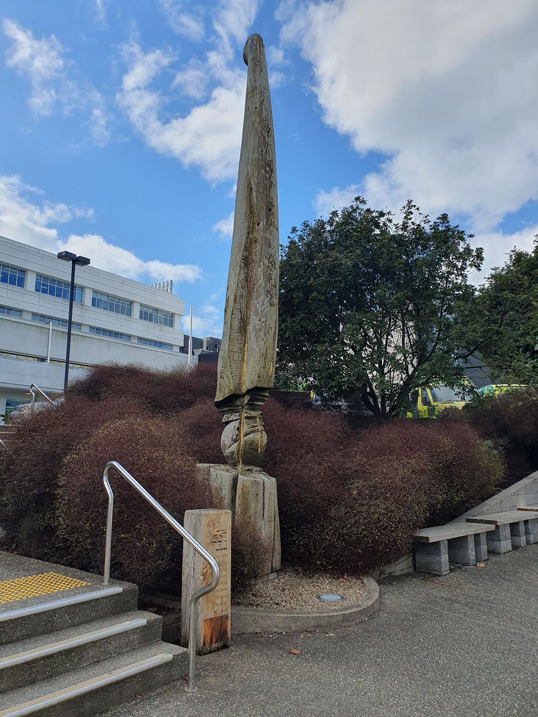 A tall carved wooden column rises from a planted bed beside steps at North Shore Hospital.