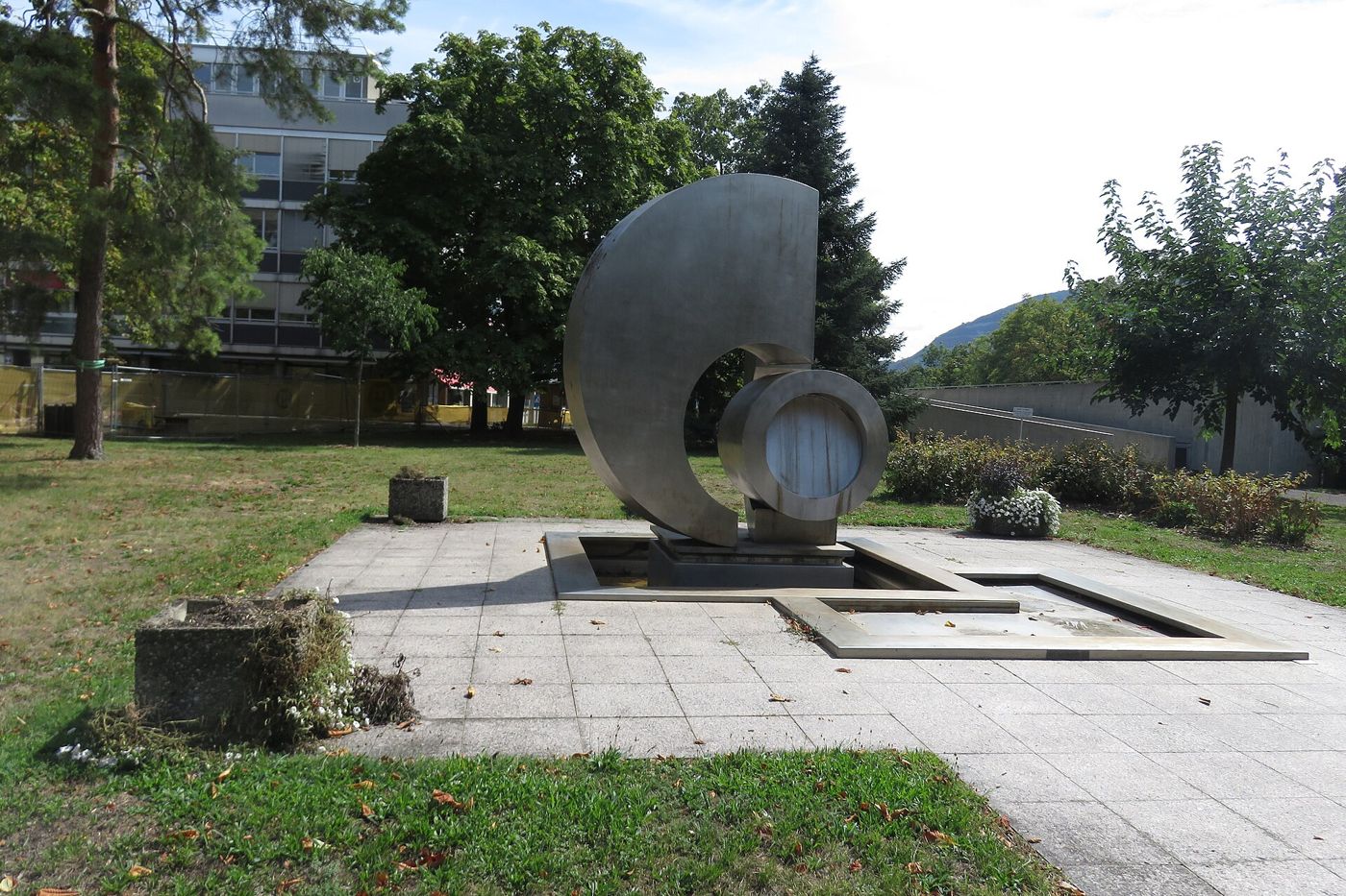A curved stainless-steel crescent sculpture stands in a paved area with a basin, surrounded by lawn and trees.