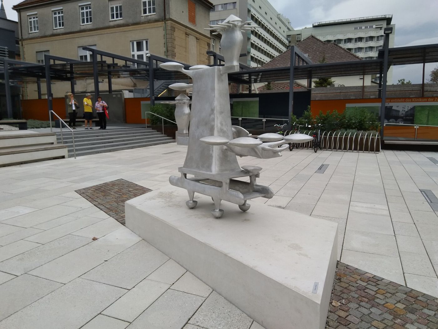 A complex silver cast-aluminium sculpture on a white plinth in a paved forecourt in front of hospital buildings.