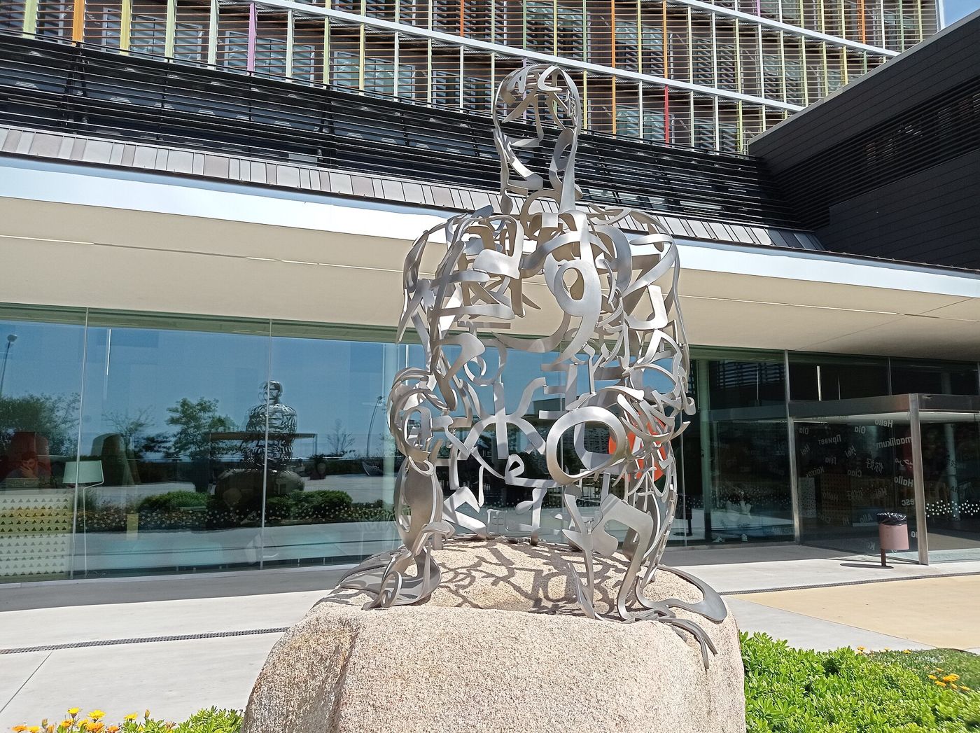 A figure made of interlaced metal letters sits on a rounded stone in front of the glass facade of Hospital Sant Joan de Déu.