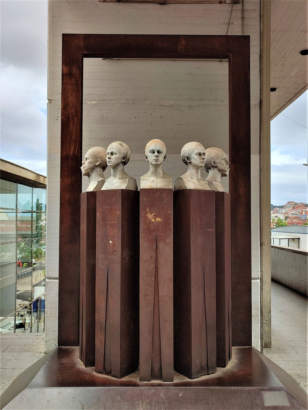 Five sculpted heads on rusted steel pillars stand inside a large rusted steel door frame at Vall d’Hebron Hospital.