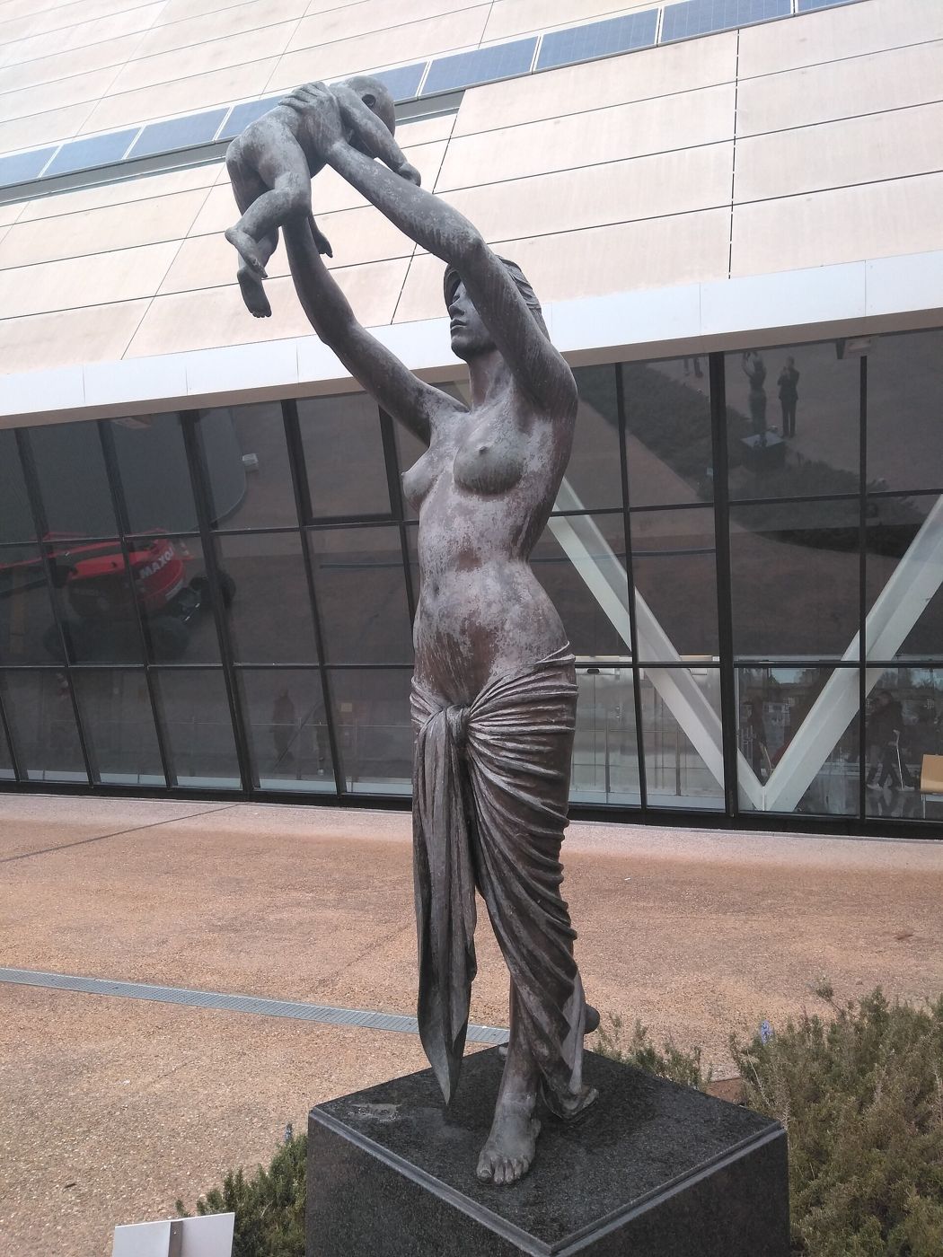 A standing female figure lifts a small child high above her head in front of a hospital building.
