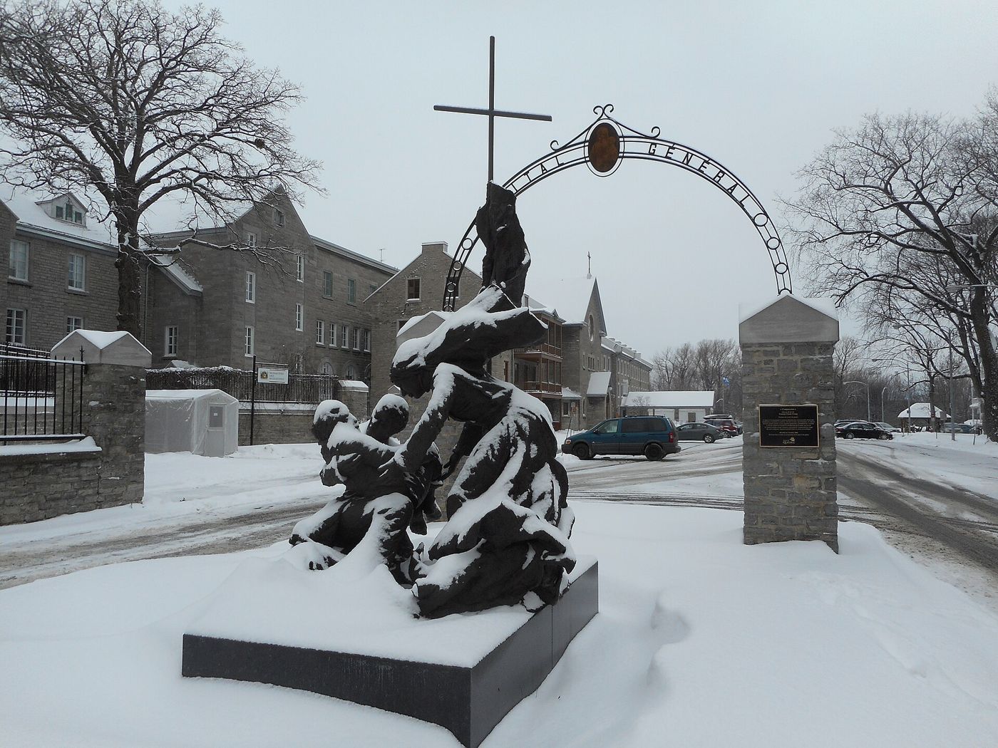 A dark sculpted group stands on a plinth before an entrance arch with a cross, under snow.