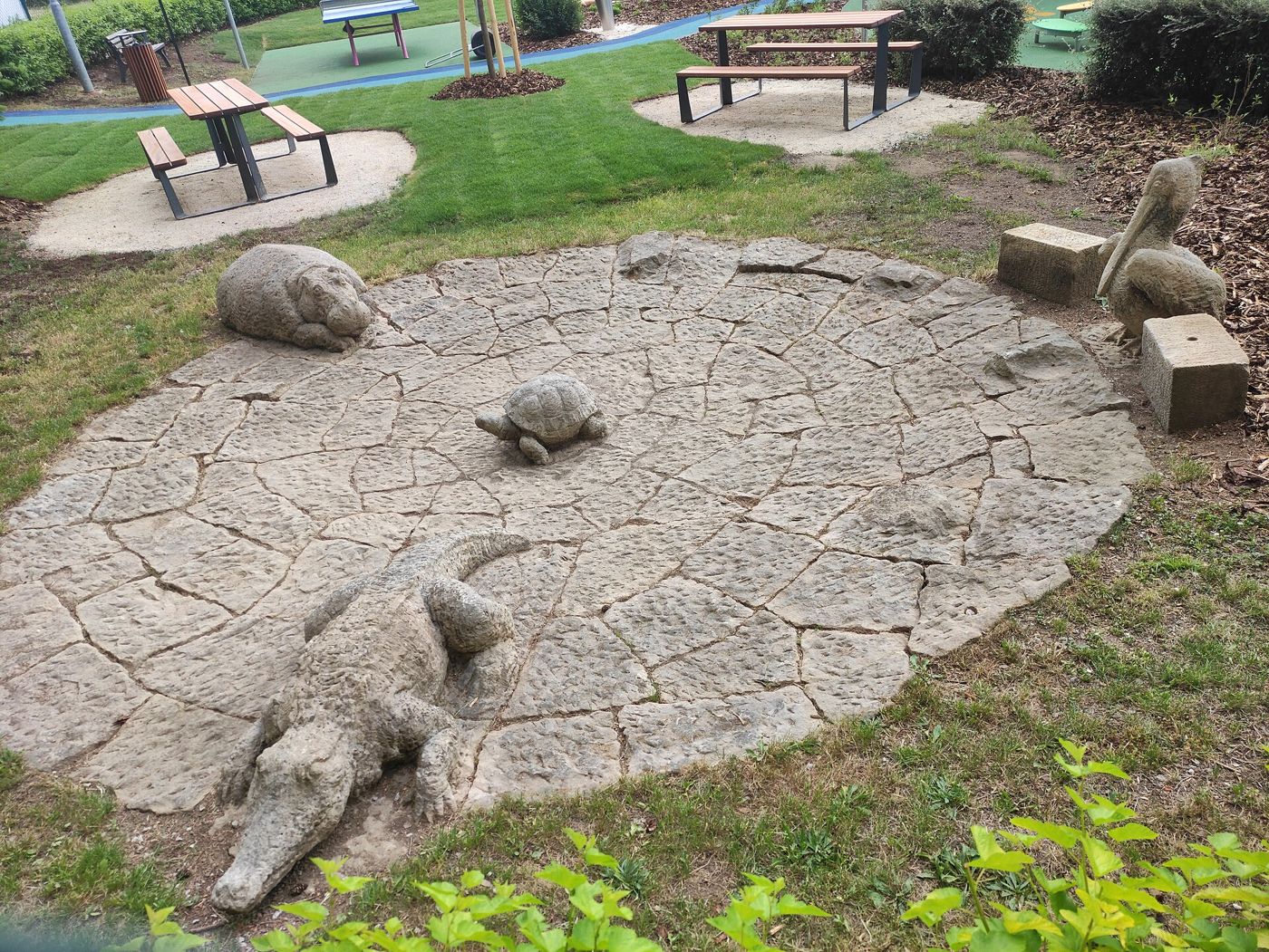 Sandstone animals, including a crocodile and a tortoise, lie on a paved circle in a garden with benches.