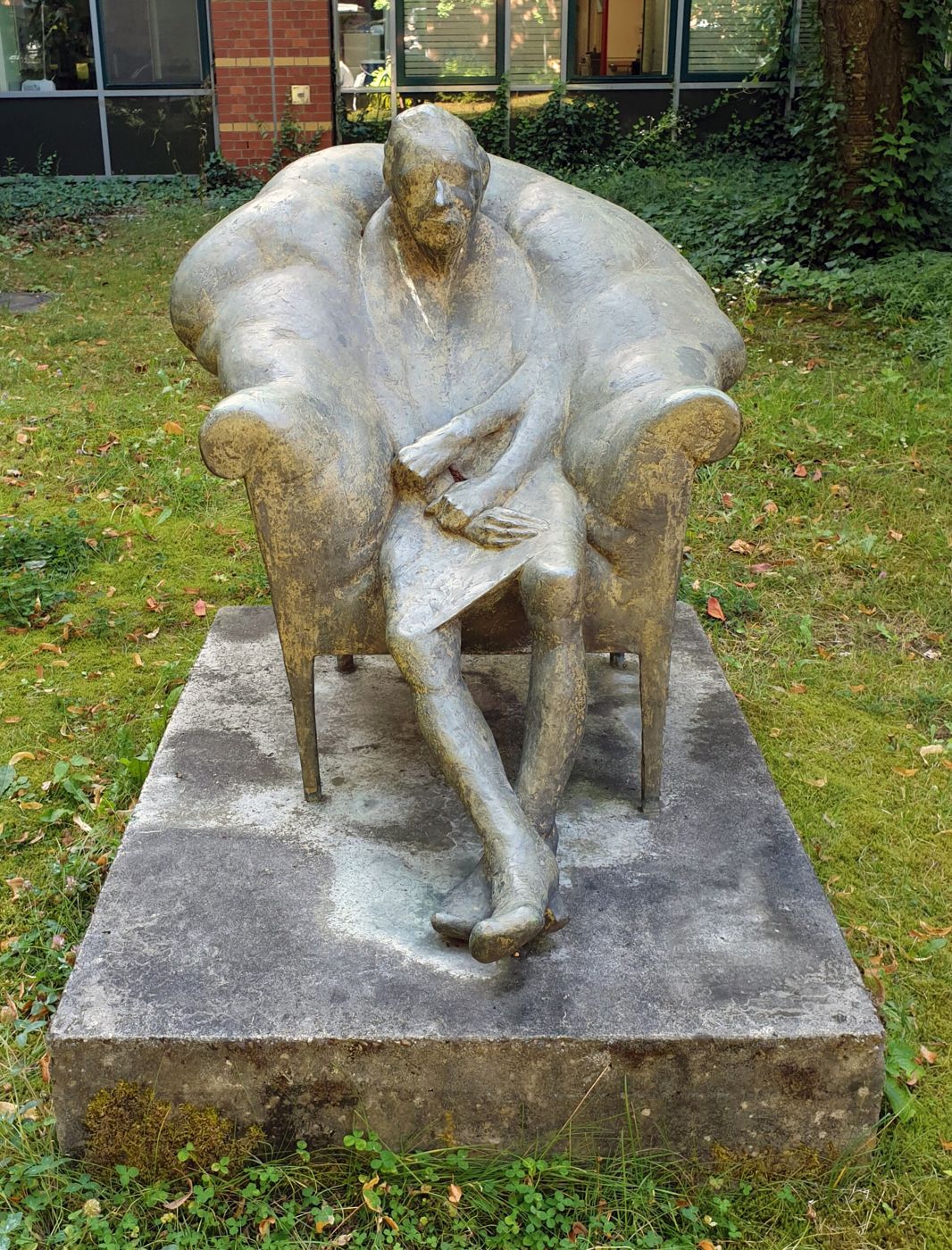 A bronze elderly woman sits in a wing chair on a stone base on a lawn.